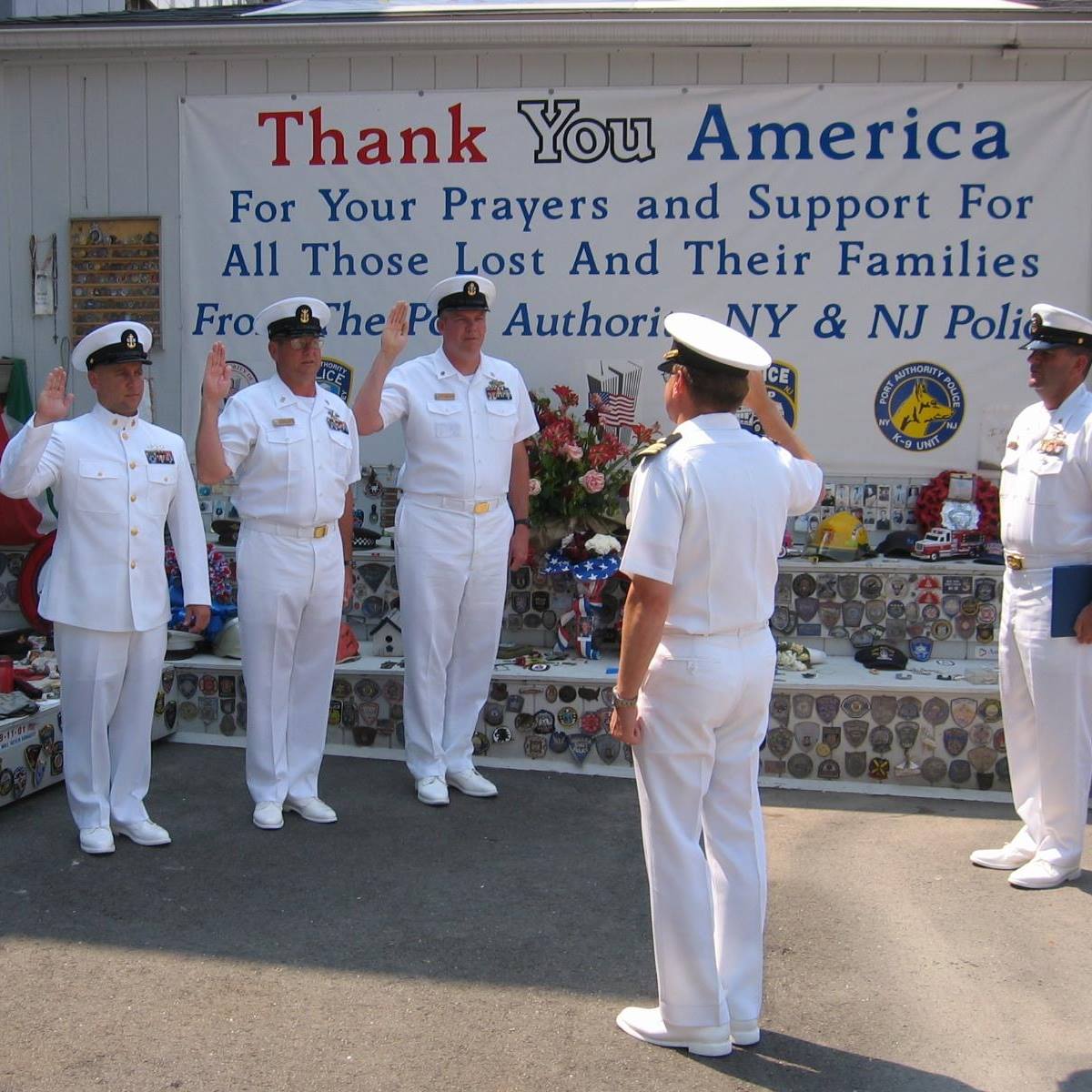 Sailors in dress whites at a ceremony in front of a “Thank You America” banner